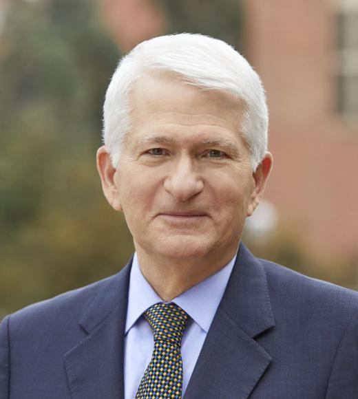 A headshot of Gene standing outside against against distant and blurry campus scenery wearing a white dress shirt, a blue suit jacket, and a tie. Gene is smiling, with hazel eyes, short white hair, and light-toned skin.
