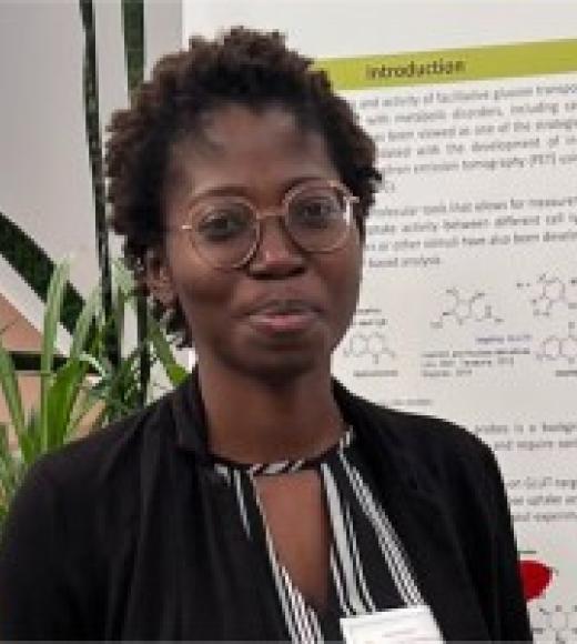 A headshot of Monica Nyansa standing in front a poster wearing a dress shirt and a black jacket. She is Smiling, with brown eyes, short black hair, dark skin, and glasses.