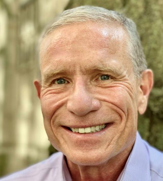 A headshot of Michael Blayney standing against distant blurry greenery. Michael is smiling big with blue-green eyes, short grey hair, and light-toned skin.