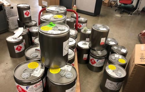 Around 30 150 gallon drums of flammable solvents stacked on the floor and on a table.