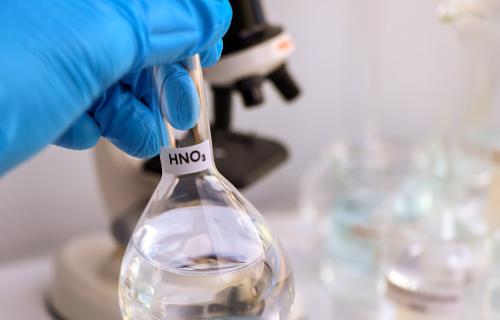 A gloved hand holding a volumetric flask with a clear liquid in it labeled as nitric acid