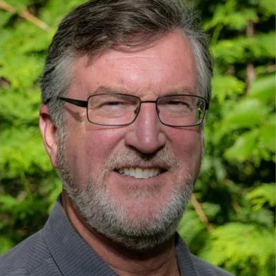 A headshot of Craig Merlic standing outside against in front of some greenery wearing a blue dress shirt. Craig is smiling, with squinted eyes in the sunlight. He has short greyed hair and a short grey beard with glasses.