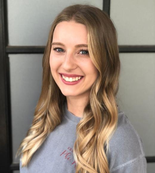 A headshot of Brayley Gattis standing against a white tiled wall. Brayley is facing to the left and smiling a big, toothy smile with big blue eyes, long blond wavy hair and light-toned skin.