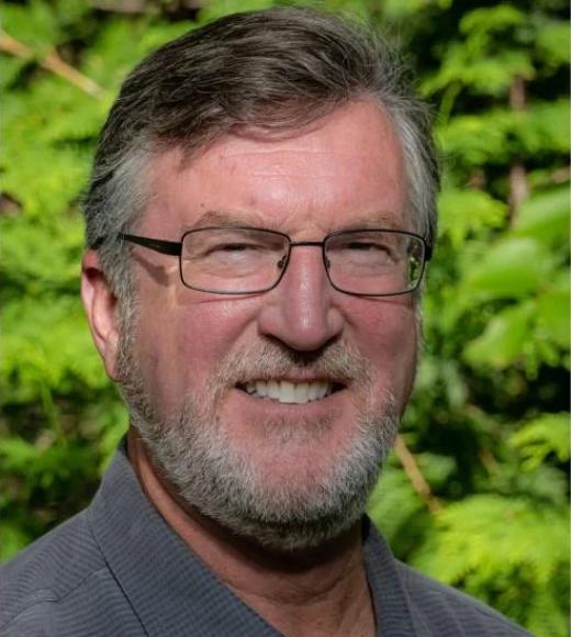 A headshot of Craig Merlic standing outside against in front of some greenery wearing a blue dress shirt. Craig is smiling, with squinted eyes in the sunlight. He has short greyed hair, a short grey beard with glasses, and light-toned skin.