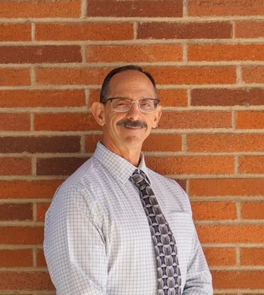 A headshot of Curtis Plotkin standing against a deeply-colored brick wall wearing a white dress shirt with a tie. Curtis is smiling, with squinted eyes. He has very short peppered hair, a thick peppered mustache, glasses, and light-toned skin.