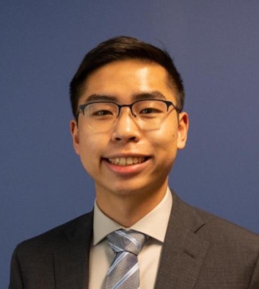 A headshot of Daniel Hu standing against a dark blue background wearing a white dress shirt, a black suit jacket, and a light blue tie. Daniel has a big smile with dark brown eyes, short black hair, glasses, and medium-toned skin.