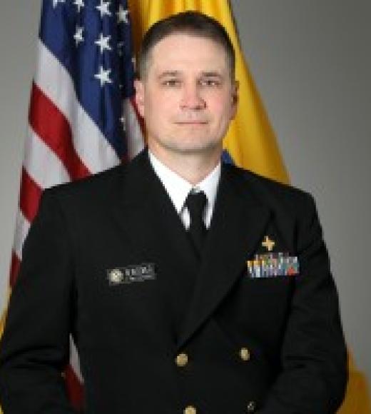 A headshot of Derek Newcomer standing against a white wall with an American flag. Derek is wearing a white dress shirt with a suit jacket and a tie, with several medals adorning his jacket. Derek is sporting a reserved smile, brown eyes, a brown buzzcut, and light-toned skin.