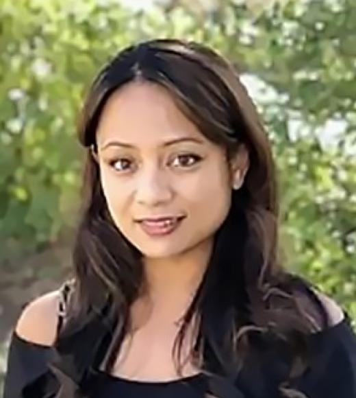 A headshot of Jannette Ducut. Janet is standing against some green foliage wearing a black off-the-shoulder shirt. Janet is smiling faintly, with big brown eyes, long brown hair, and medium-toned skin.