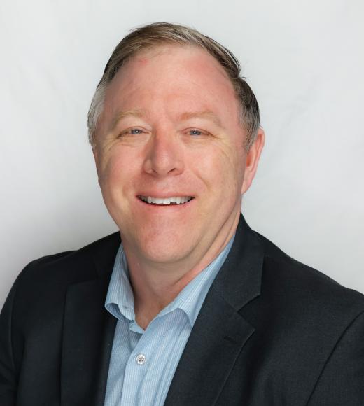 A headshot of Ken Smith standing against a white background wearing a light blue dress shirt and black suit jacket. Ken is smiling with blue, squinty eyes, short peppered hair, and light-toned skin.