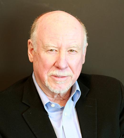 A headshot of Tom Krause standing against a dark olive green background wearing a light blue dress shirt and black suit jacket. Tom is not smiling, with greenish eyes, white hair, a bald head with white hair on the sides, and light-toned skin.