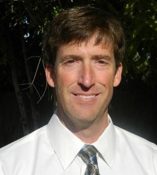 A headshot of Brent Cooley wearing a white dress shirt and tie, standing against foliage. Brent has medium-dark brown hair, light skin, and brown eyes. He is smiling and squinting slightly in the sun.