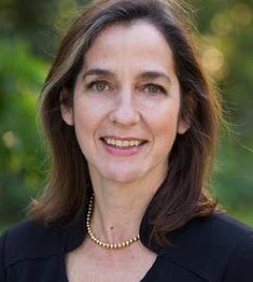 A headshot of Camille Peres standing outside against some distant blurry greenery wearing a dark blue shirt. Camille is smiling, with hazel-green eyes, shoulder-length brown hair, and light-toned skin.