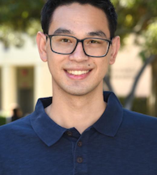 A headshot of Ethan Raffman. Ethan is standing in front of distant and blurry outdoor scenery wearing a dark blue polo shirt. Ethan is smiling with dark brown eyes, short black hair, glasses, and light-toned skin